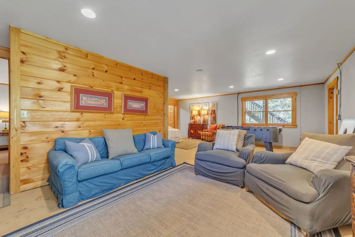 A welcoming family room with blue couches set against a wood-paneled wall, a patterned area rug, framed artwork, and ample windows inviting in natural light. The space is arranged for socializing, reading, and playing games, creating a cozy and homely atmosphere ideally suited for gatherings.