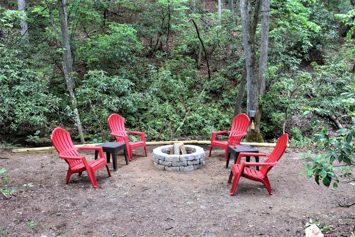 A cozy outdoor gathering space featuring red Adirondack chairs arranged around a stone fire pit amidst lush greenery. The setting invites storytelling, s'mores, and stargazing under the open sky.
