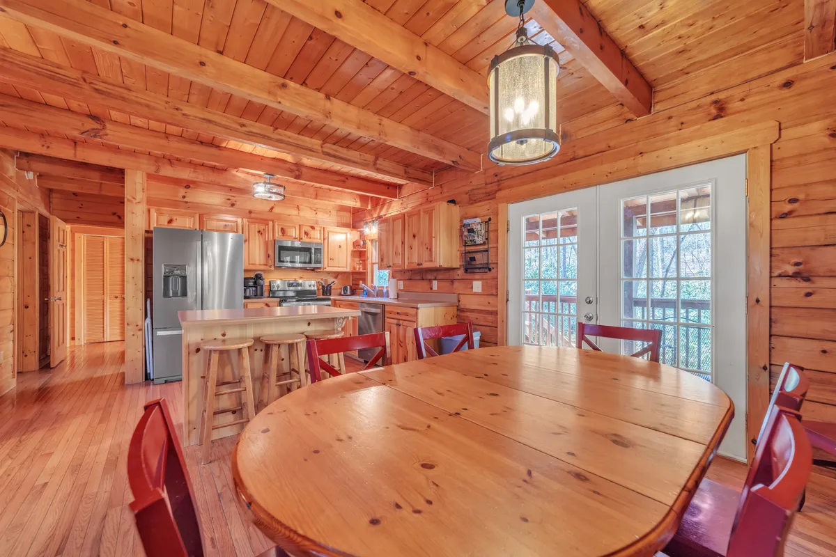 An expansive wooden dining table takes center stage in an airy, inviting room, with kitchen appliances and wooden ceiling beams visible. Double doors open to the outdoors, highlighting the seamless flow between interior dining and nature.