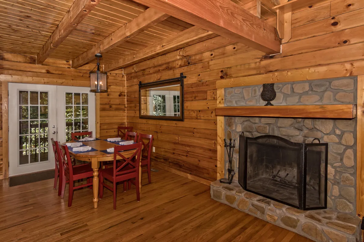 A charming dining area with a wooden table, a crackling fireplace, exposed wooden beams, and French doors that hint at access to the outdoors. The setup suggests intimate meals and relaxing by the fire.