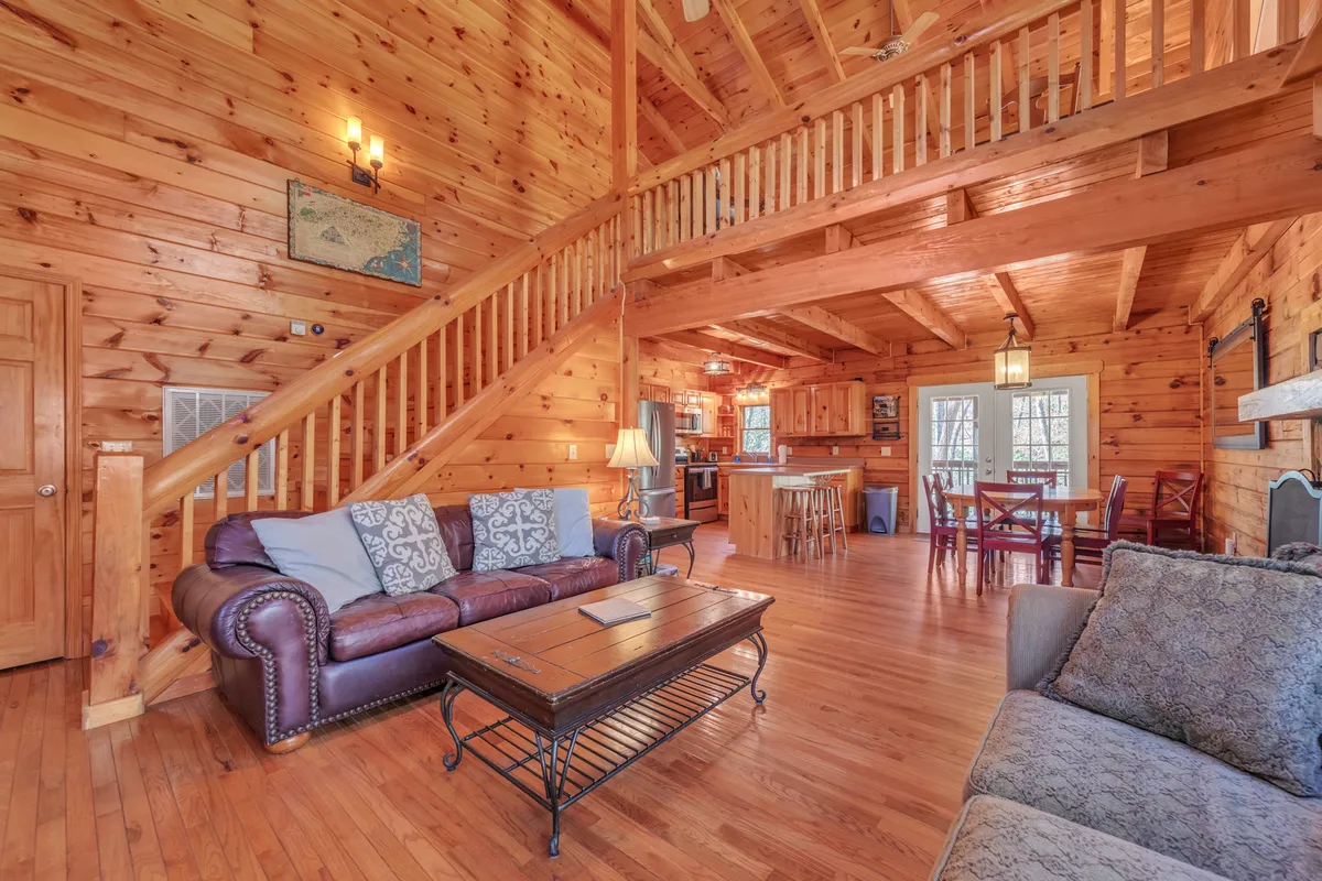 A spacious open interior featuring a wooden staircase, comfortable sofas, a coffee table, and a view of both the kitchen and dining area. The room is designed for family gatherings, relaxation, and entertaining, with visible open-beam construction adding rustic charm.