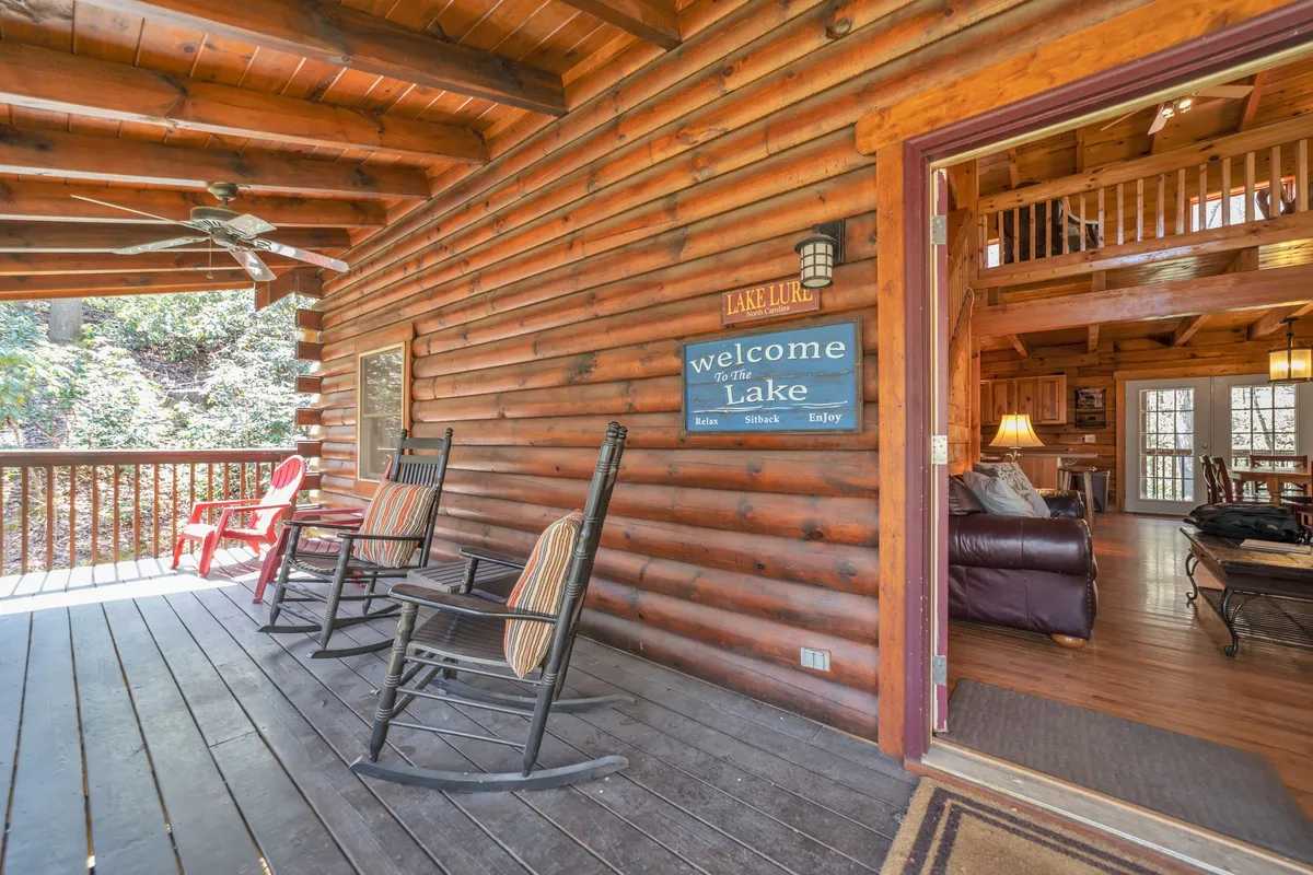 This image captures the welcoming exterior of the cabin, featuring inviting rocking chairs, a rustic welcome sign, wooden beams, and a beautiful view of the lake. Guests can be seen relaxing on the porch, enjoying the natural scenery and atmosphere.