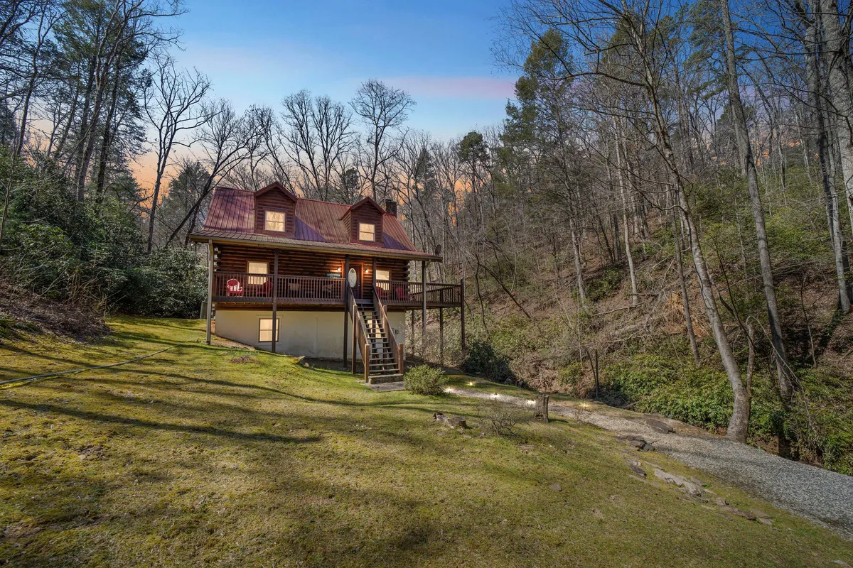 The image showcases a charming log cabin nestled among towering trees, with a spacious wooden deck perfect for relaxing. A moss-lined pathway leads guests through the lush woods towards the inviting cabin, hinting at the tranquil natural surroundings and the adventure that awaits just steps from the porch.