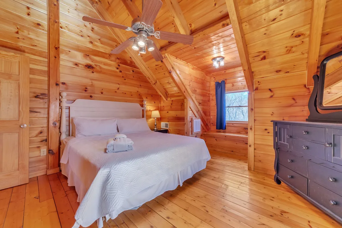 A serenely styled bedroom featuring a queen bed, wooden ceiling and walls, a dresser, a bedside lamp, and a window with curtains framing views of the natural landscape. The mood is peaceful and inviting, perfect for restful getaways.