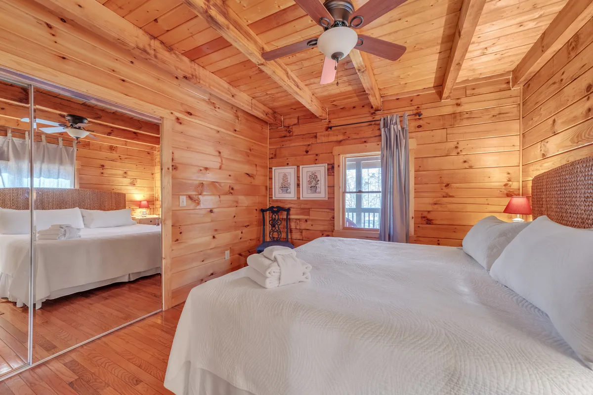 A serene bedroom scene featuring a bed with crisp white bedding, a mirrored wardrobe, a nightstand with a lamp, and framed artwork on the walls. Natural light pours in, accentuating the cozy atmosphere created by the wooden ceiling and soft furnishings.