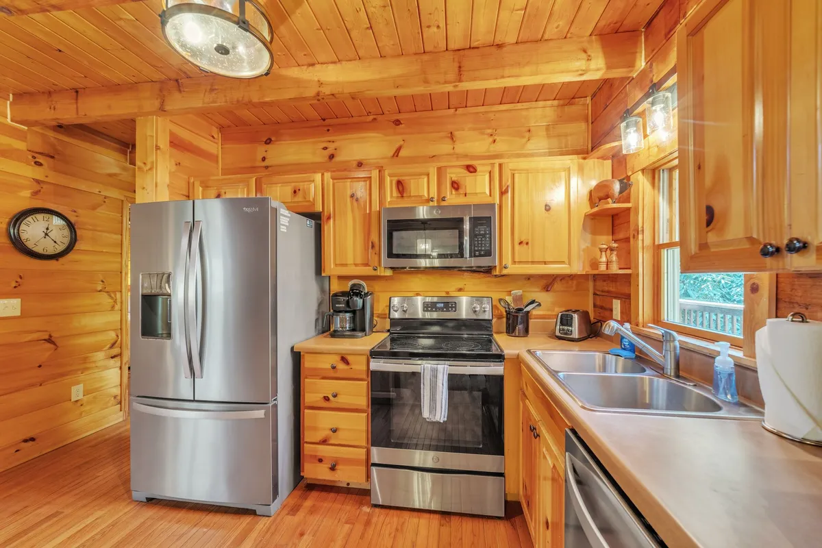 A cozy, rustic kitchen space featuring a stainless steel refrigerator and oven, wooden cabinets, and a sink set beneath a window that offers views of the surrounding forest. The space is warm and inviting, ideal for preparing meals and gathering together.