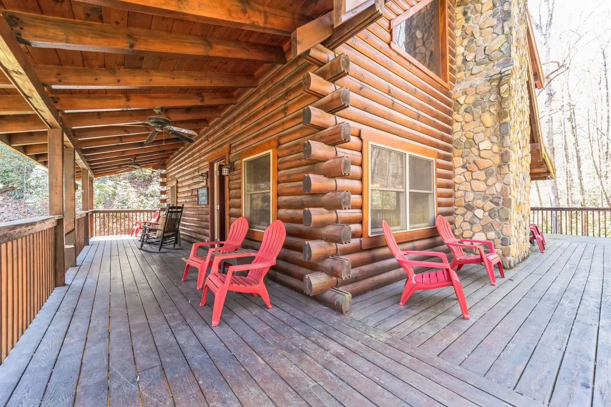 The image shows a classic log cabin structure with red Adirondack chairs arranged on a porch, set against a stone wall. This outdoor seating area encourages relaxation, gathering, and enjoying the peace of the natural surroundings.