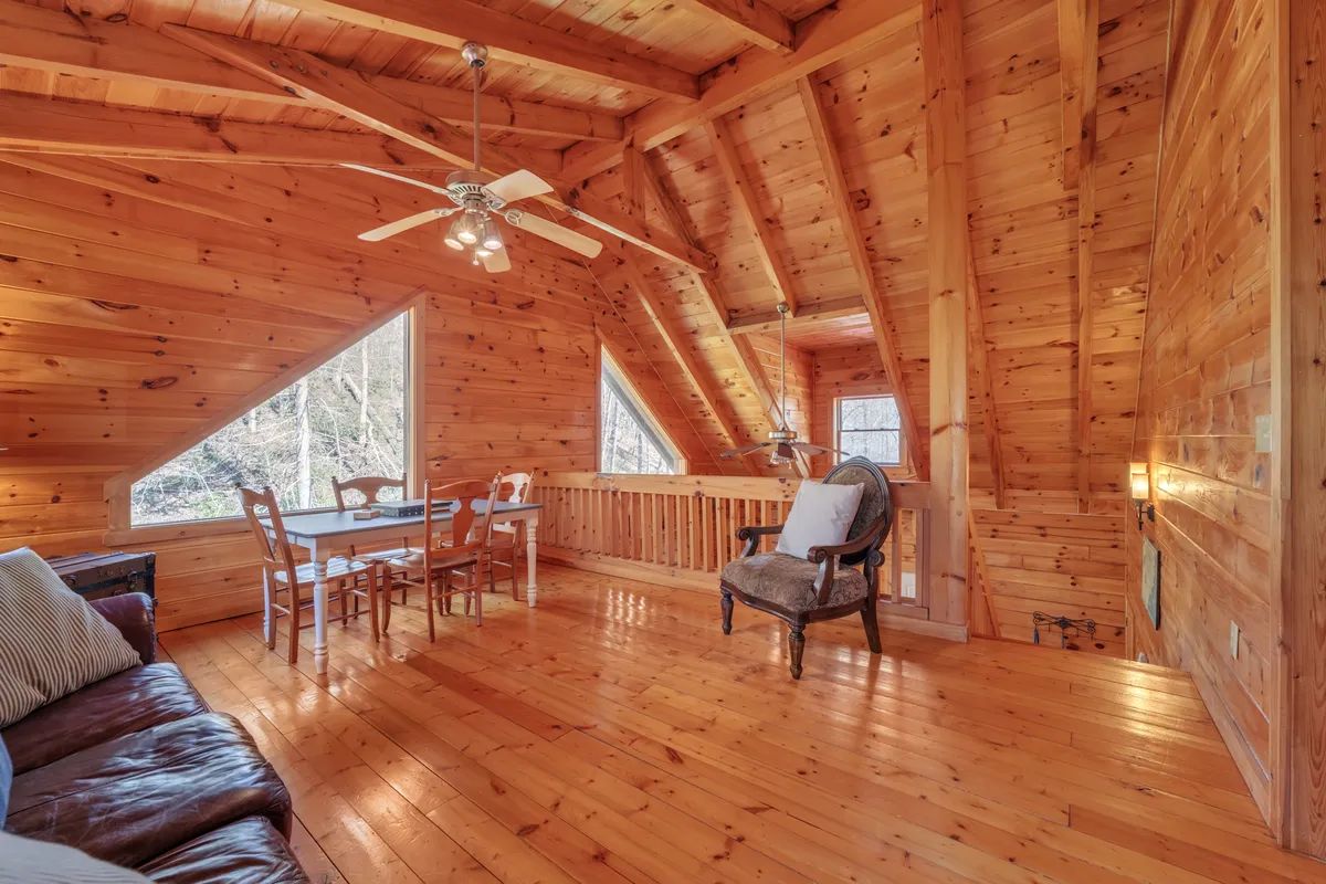 A warm, open living space with rustic wooden beams, large windows offering glimpses of the outdoors, a dining table set for two, and a cozy sofa and chair. The soft lighting and visible sunset glow through the windows evoke a romantic, tranquil ambiance.
