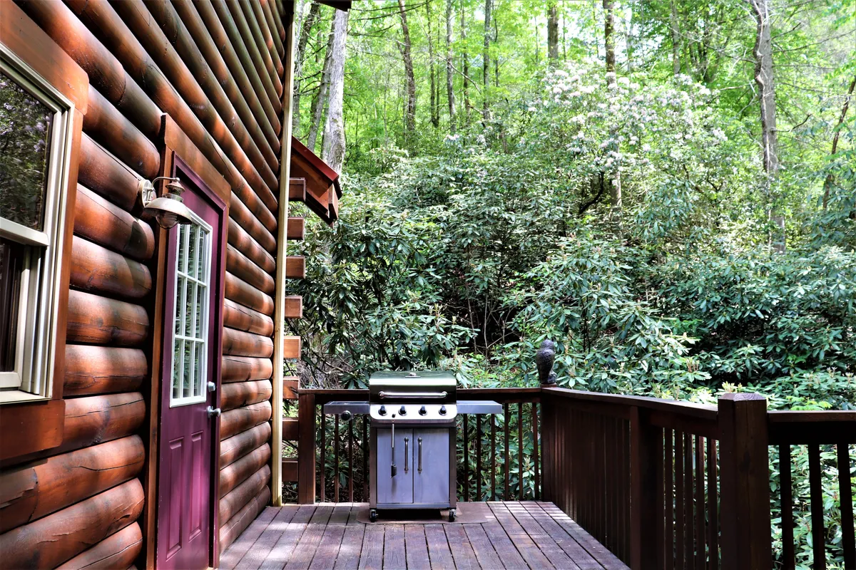 The deck of the log cabin features a grill, with lush greenery providing a scenic backdrop. The outdoor setup is perfect for grilling and enjoying meals al fresco while surrounded by nature.